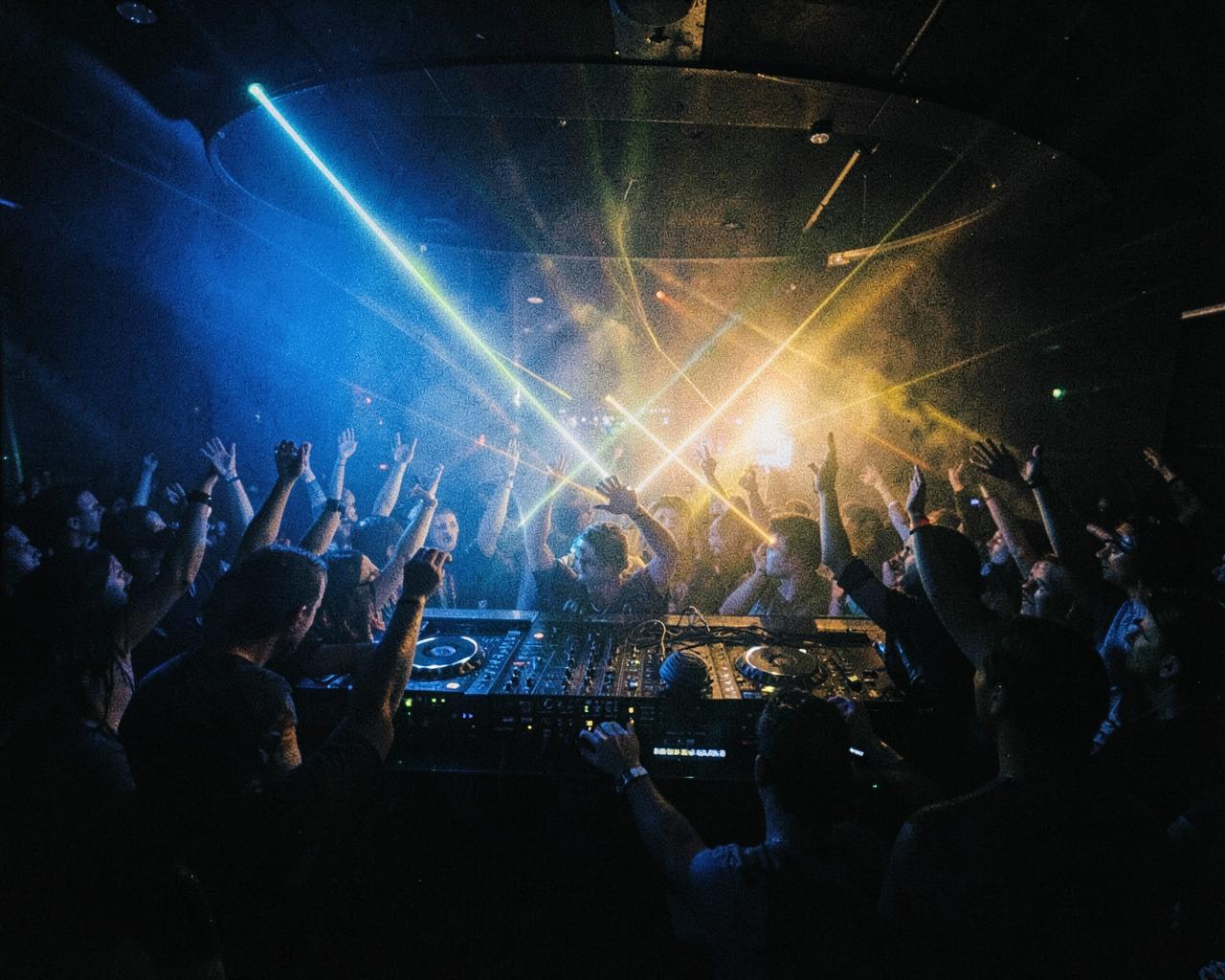 Crowd with hands raised in front of a DJ booth under blue and gold lights